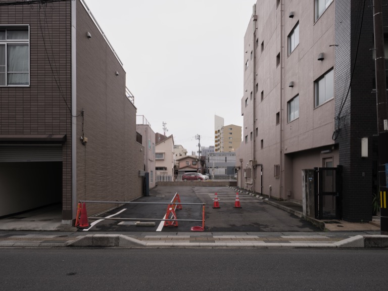 An empty space for parking can be found between houses and buildings. There are no cars parked, and the ground surface is nearly dry, with a slight dampness probably resulting from recent rain.